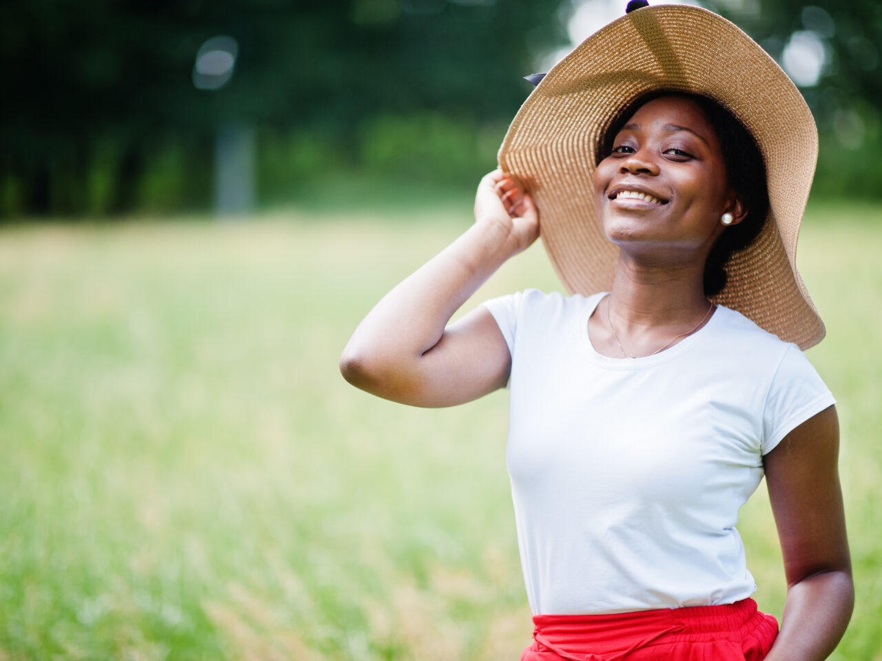 Portrait of gorgeous african american woman 20s wear in summer h Portrait of gorgeous african american woman 20s wear in summer hat, red pants and white t-shirt, posing at green grass in park.